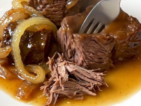 A close-up of a slow cooker filled with tender chunks of chuck roast and red onions in a rich, glossy hot honey sauce.