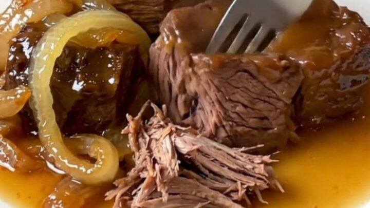 A close-up of a slow cooker filled with tender chunks of chuck roast and red onions in a rich, glossy hot honey sauce.
