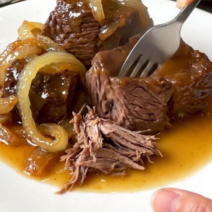 A close-up of a slow cooker filled with tender chunks of chuck roast and red onions in a rich, glossy hot honey sauce.