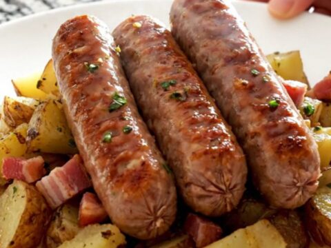 A white plate topped with braised bratwurst, tender red potato rounds, and crispy bacon in a tangy sauce.