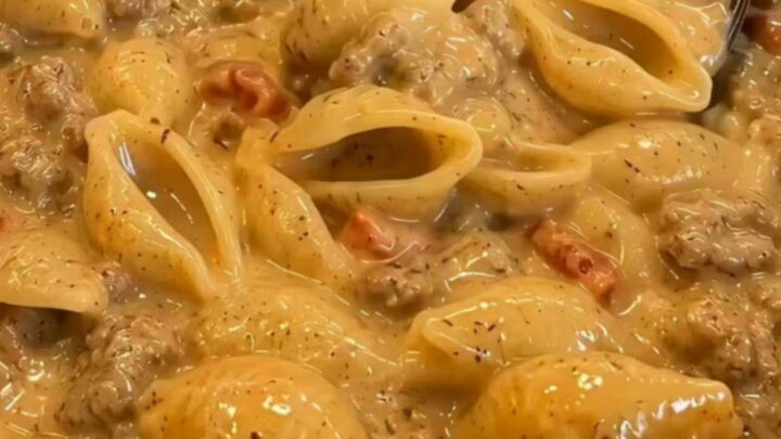 A wooden spoon lifting glossy shell pasta dripping in a rich, seasoned taco-cheese sauce from a skillet.