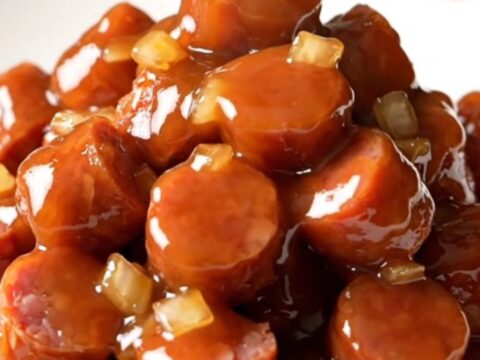 A slow cooker filled with golden-brown kielbasa coins coated in a thick, shiny honey barbecue sauce with bits of onion.