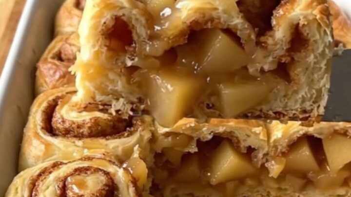 A golden, baked apple pie cinnamon roll dish in a white ceramic pan on a wooden counter.