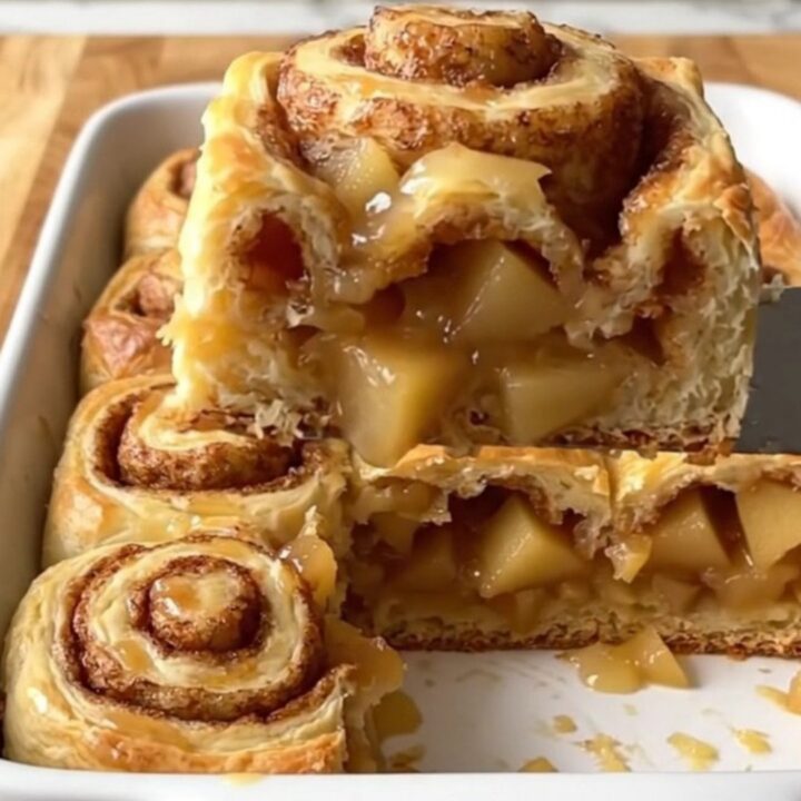 A golden, baked apple pie cinnamon roll dish in a white ceramic pan on a wooden counter.