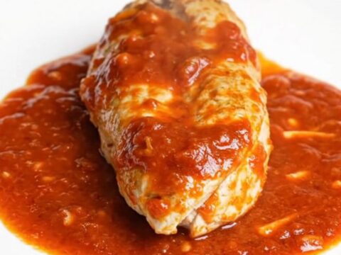 A serving spoon lifting a tender chicken breast coated in thick, cheesy marinara sauce from a black slow cooker.