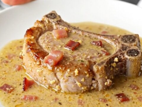 A close-up of a juicy, braised pork chop covered in a thick, creamy bacon-flecked gravy in a slow cooker.