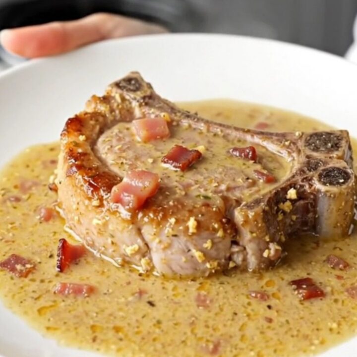 A close-up of a juicy, braised pork chop covered in a thick, creamy bacon-flecked gravy in a slow cooker.
