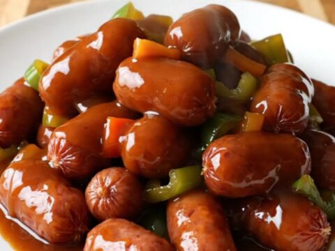 A stainless steel slow cooker filled with glazed mini smoked sausages and diced red peppers.