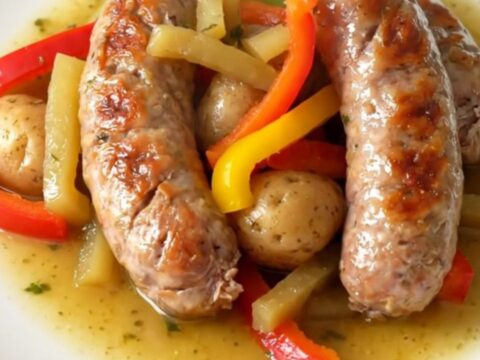 A close-up of a slow cooker filled with Italian sausage, bell pepper strips, and halved baby potatoes in a savory herb broth.