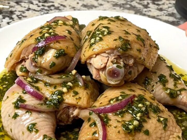 A close-up of tender braised chicken thighs in a vibrant green chimichurri sauce with red onion slices in a slow cooker.