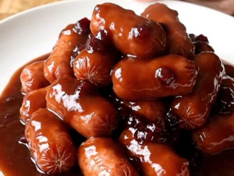 A serving spoon scoops plump mini smoked sausages coated in a thick, dark red-brown BBQ sauce from a slow cooker.