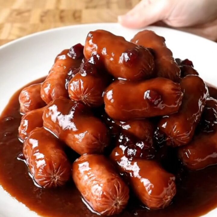 A serving spoon scoops plump mini smoked sausages coated in a thick, dark red-brown BBQ sauce from a slow cooker.