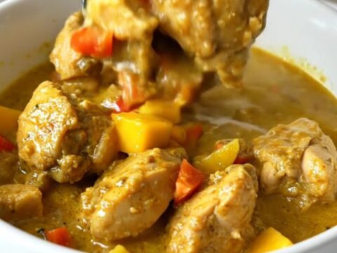 A top-down view of a slow cooker filled with chicken, mango, and red bell pepper in a golden curry sauce.