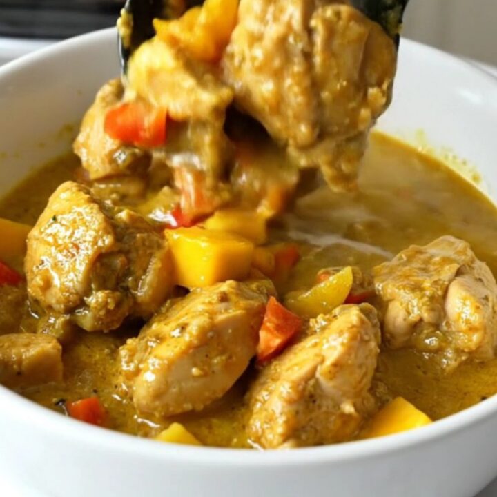 A top-down view of a slow cooker filled with chicken, mango, and red bell pepper in a golden curry sauce.