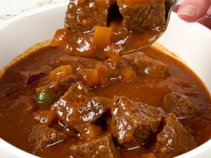 A bowl of tender slow-cooked carne guisada with a rich tomato gravy and visible pieces of beef and peppers.