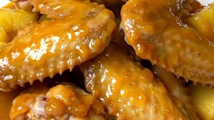 A top-down view of raw chicken wings and pineapple chunks in a black slow cooker insert on a granite countertop.