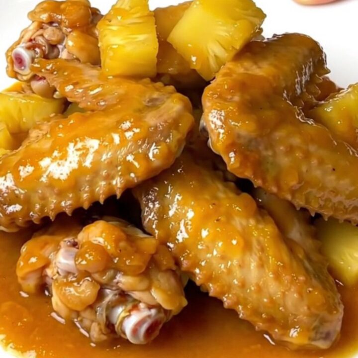A top-down view of raw chicken wings and pineapple chunks in a black slow cooker insert on a granite countertop.