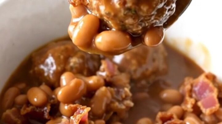 A close-up of a white bowl filled with cowboy meatballs, pinto beans, and bacon in a rich barbecue sauce.