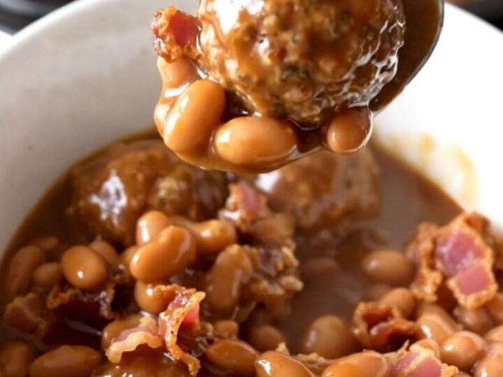A close-up of a white bowl filled with cowboy meatballs, pinto beans, and bacon in a rich barbecue sauce.