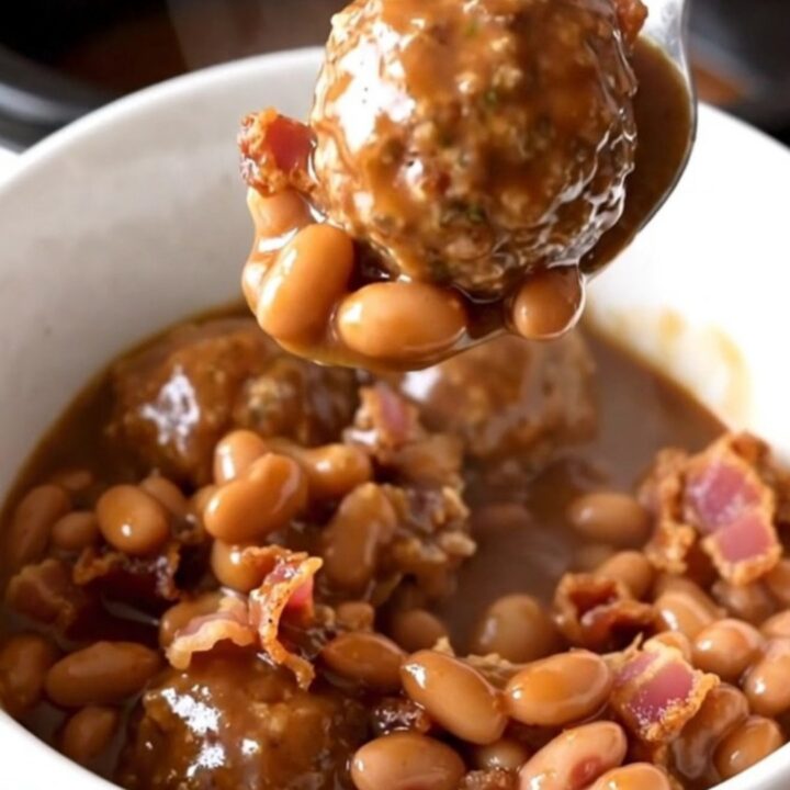 A close-up of a white bowl filled with cowboy meatballs, pinto beans, and bacon in a rich barbecue sauce.