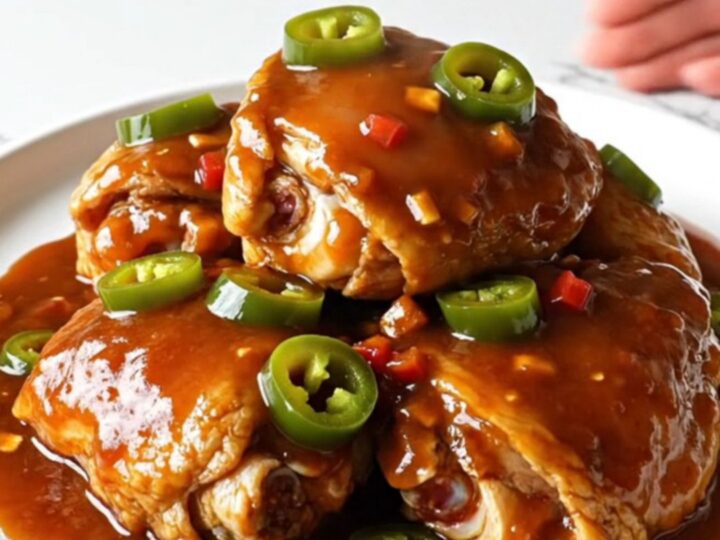 A serving spoon lifting a tender chicken thigh coated in a thick, glossy red-brown BBQ sauce with visible jalapeno rings.