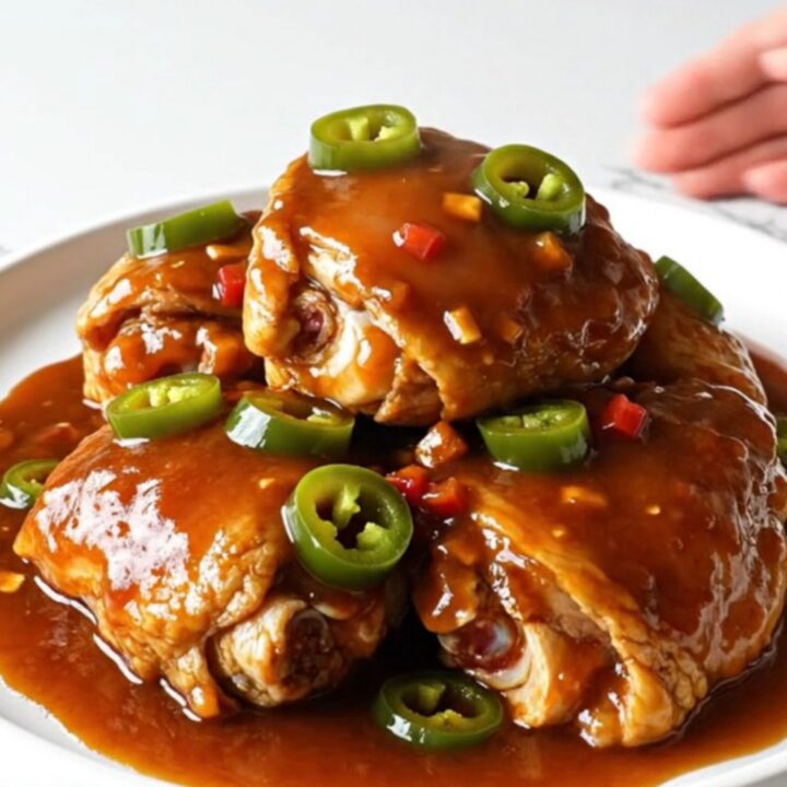 A serving spoon lifting a tender chicken thigh coated in a thick, glossy red-brown BBQ sauce with visible jalapeno rings.