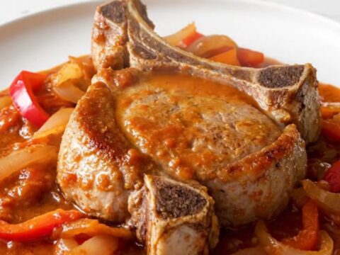 A close-up of a tender bone-in pork chop topped with a rich tomato, pepper, and onion sauce on a white plate.