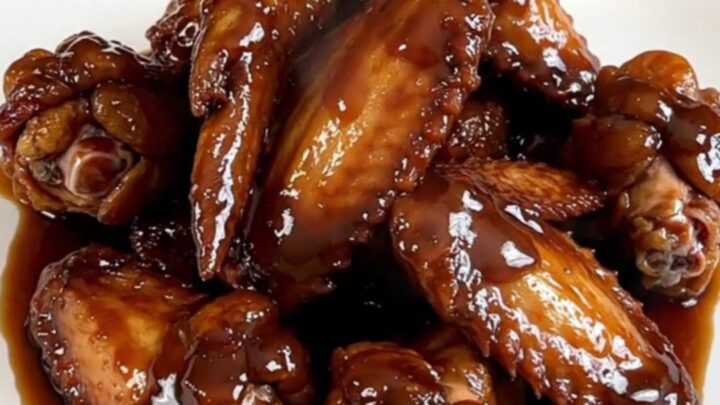 A close-up of dark, glazed chicken wings on a white plate with a glossy, thick soy molasses sauce and light steam.