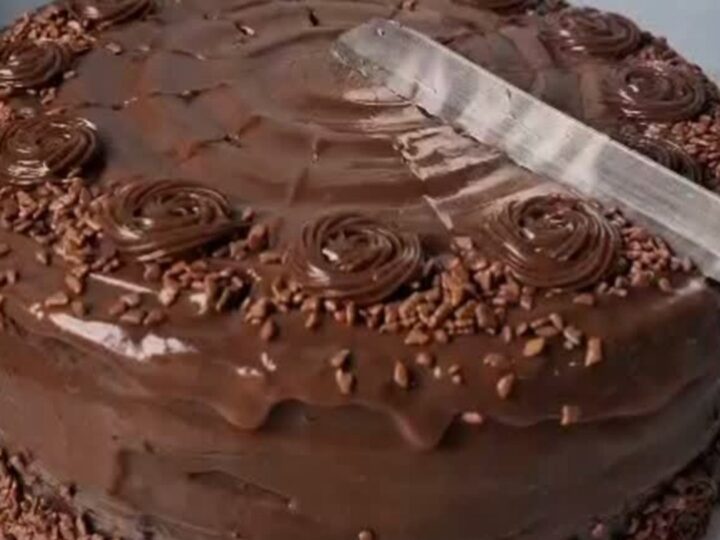 A decadent chocolate fudge cake with glossy ganache and piped rosettes on a white scalloped stand.