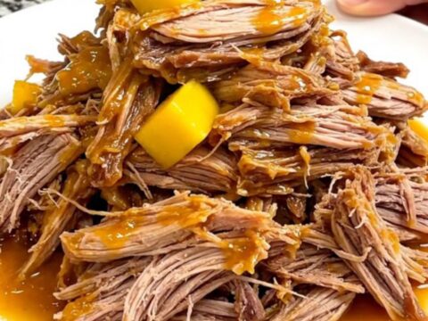 A close-up of tender mango habanero pulled pork served on a white plate with visible strands of meat and soft mango pieces.