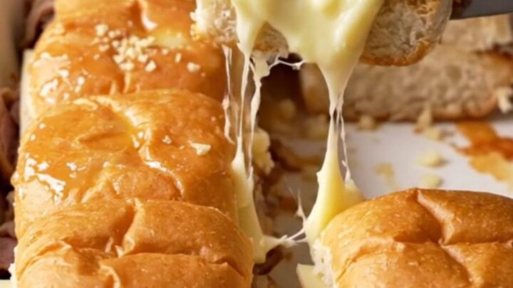 A close-up of golden-brown French dip sliders in a white baking dish with melted provolone and roast beef.