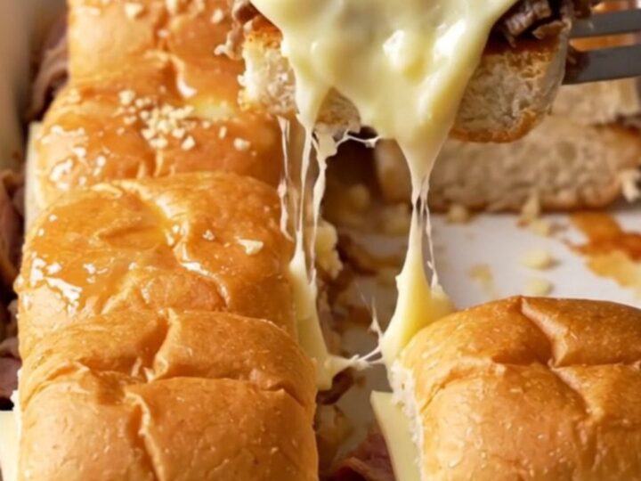 A close-up of golden-brown French dip sliders in a white baking dish with melted provolone and roast beef.