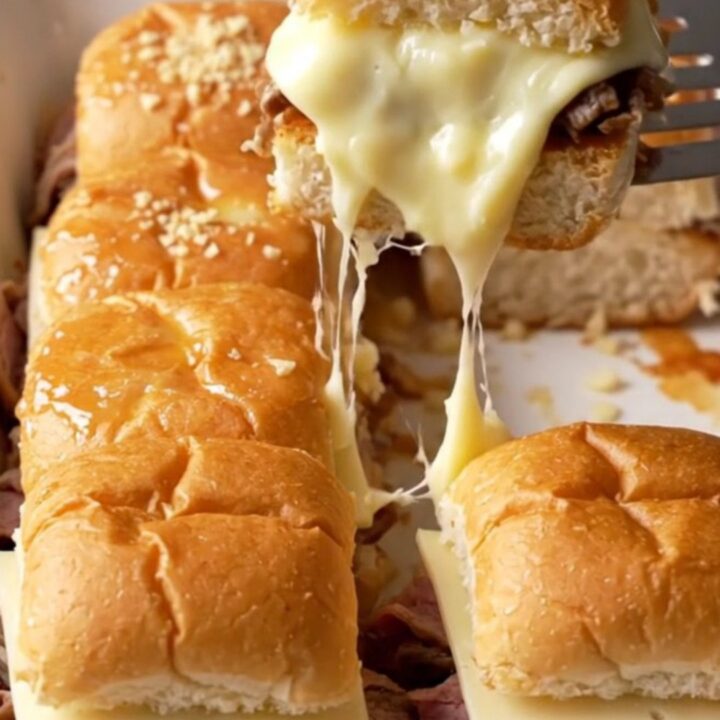 A close-up of golden-brown French dip sliders in a white baking dish with melted provolone and roast beef.