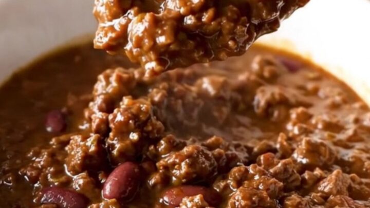 A close-up of a bowl filled with rich, dark brown beef and bean chili, garnished with a sprinkle of fresh herbs.