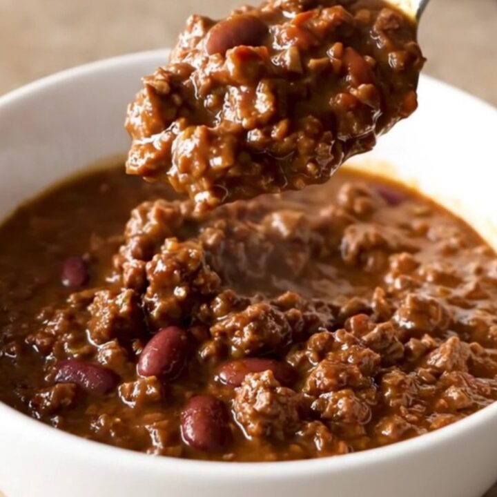 A close-up of a bowl filled with rich, dark brown beef and bean chili, garnished with a sprinkle of fresh herbs.