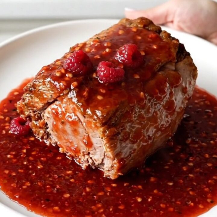 A close-up of a slow-cooked pork tenderloin smothered in a thick, glossy red raspberry pepper sauce.