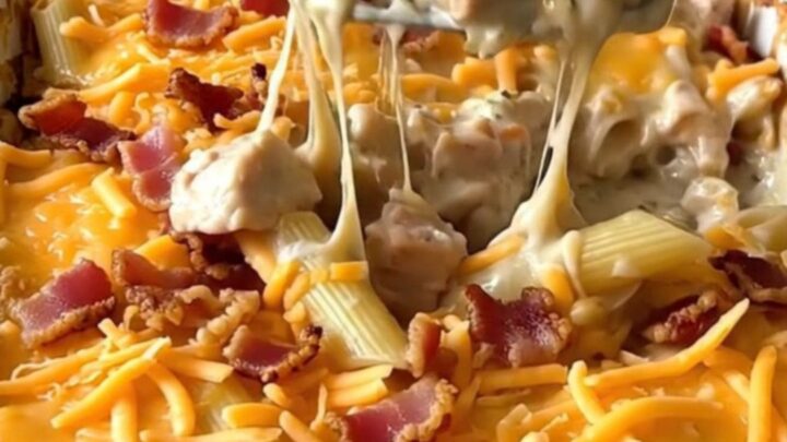 A top-down view of a rectangular baking dish filled with raw penne, chicken pieces, ranch sauce, shredded cheddar, and bacon.