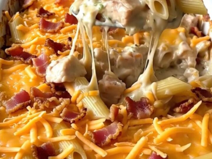 A top-down view of a rectangular baking dish filled with raw penne, chicken pieces, ranch sauce, shredded cheddar, and bacon.