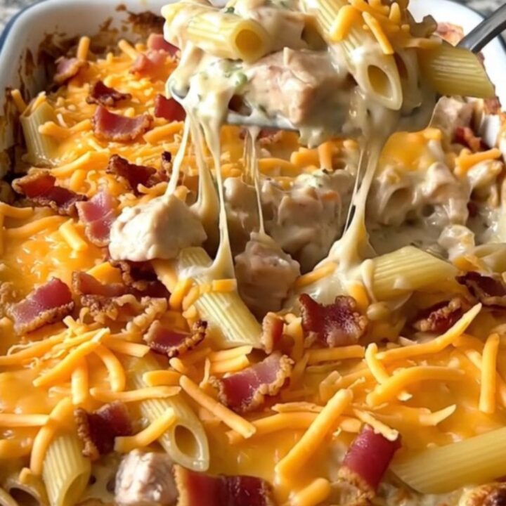 A top-down view of a rectangular baking dish filled with raw penne, chicken pieces, ranch sauce, shredded cheddar, and bacon.