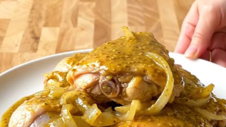 A stainless steel slow cooker filled with bone-in chicken thighs, sliced onions, and golden mustard barbecue sauce.