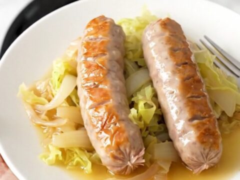 A top-down view of six raw bratwurst links arranged in a slow cooker with cabbage wedges and onions.