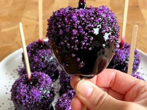 A row of glossy black poison candy apples with purple sugar sprinkles on a wooden countertop.