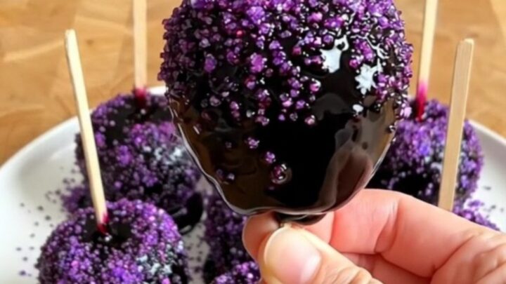 A row of glossy black poison candy apples with purple sugar sprinkles on a wooden countertop.