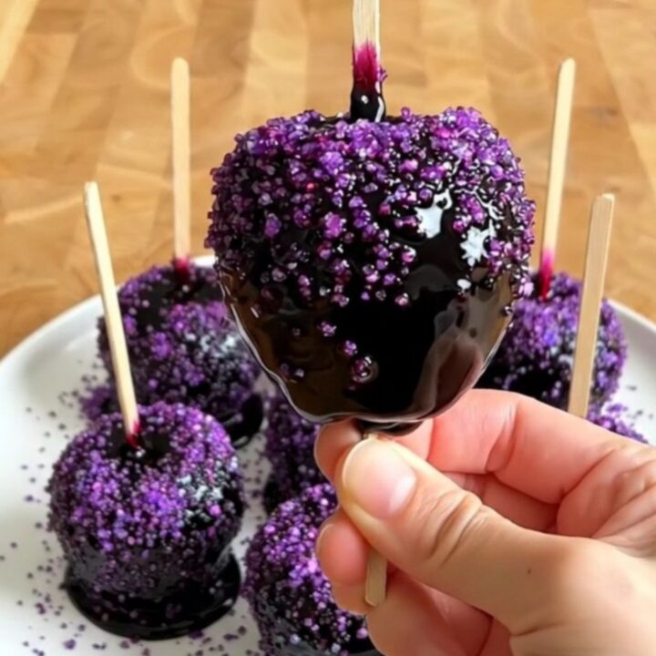 A row of glossy black poison candy apples with purple sugar sprinkles on a wooden countertop.
