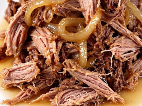 A close-up of tender, shredded vinegar pepper pulled pork in a cream-colored stoneware slow cooker.