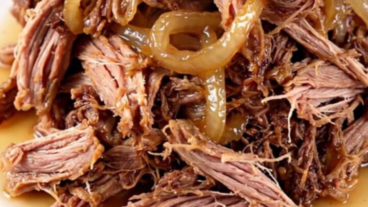 A close-up of tender, shredded vinegar pepper pulled pork in a cream-colored stoneware slow cooker.