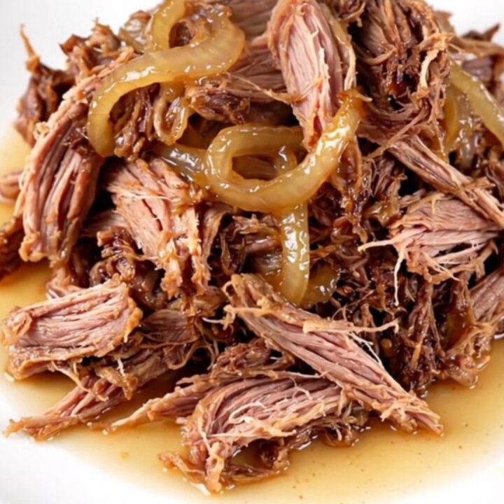 A close-up of tender, shredded vinegar pepper pulled pork in a cream-colored stoneware slow cooker.