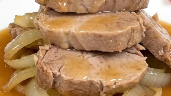 Slow cooker honey ginger pork tenderloin served on a white plate with soft onions and sauce.