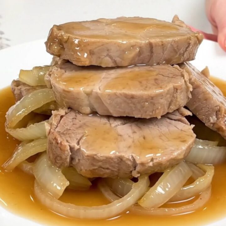 Slow cooker honey ginger pork tenderloin served on a white plate with soft onions and sauce.