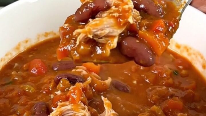 A top-down view of a black slow cooker filled with a steaming, hearty Cajun turkey chili with beans.
